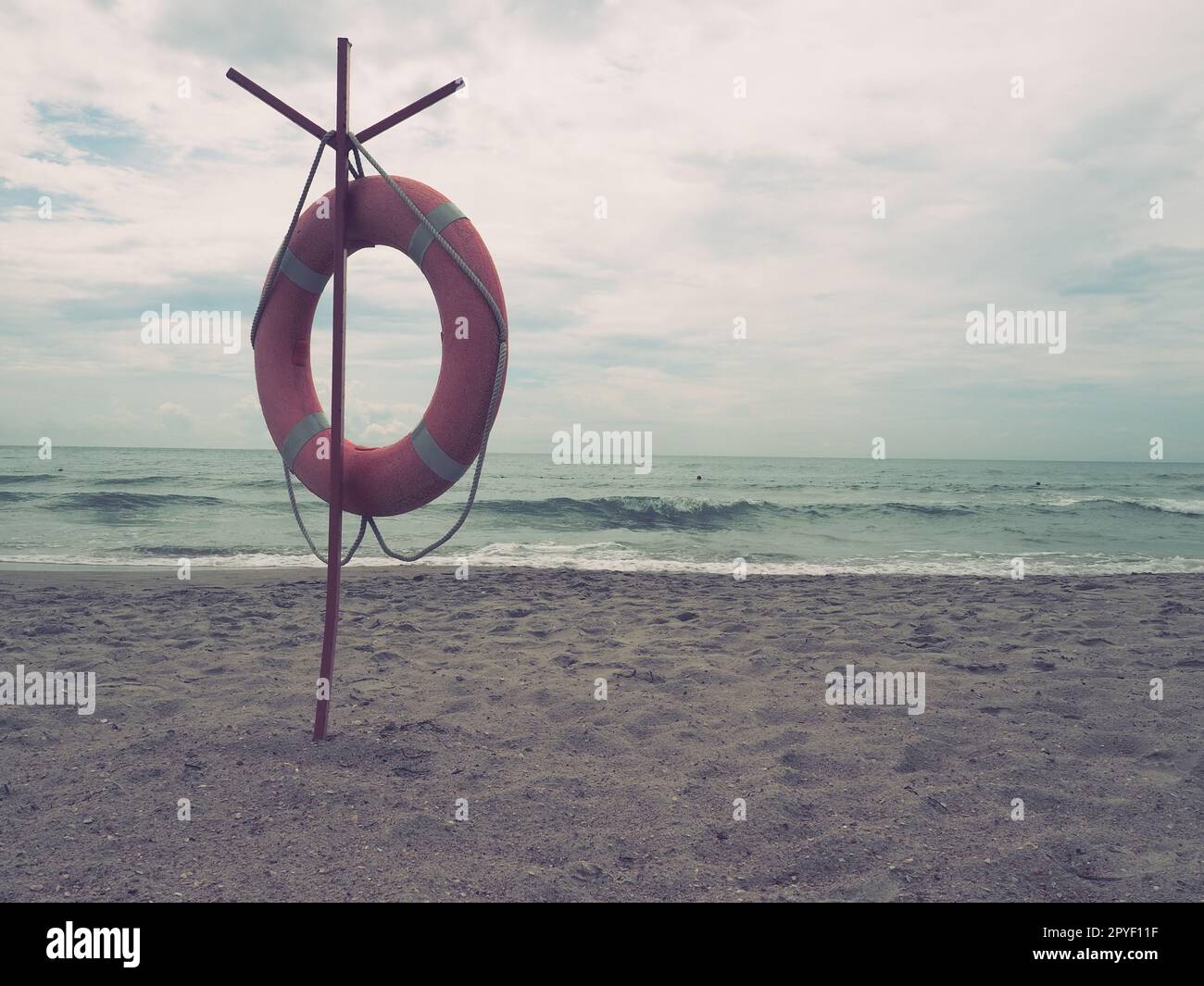 Lifebuoy on a sandy beach. Orange circle on a pole to rescue people drowning in the sea. Rescue point on the shore. Sky and sea in the background. Retro vintage photography. Nostalgic sad mood Stock Photo