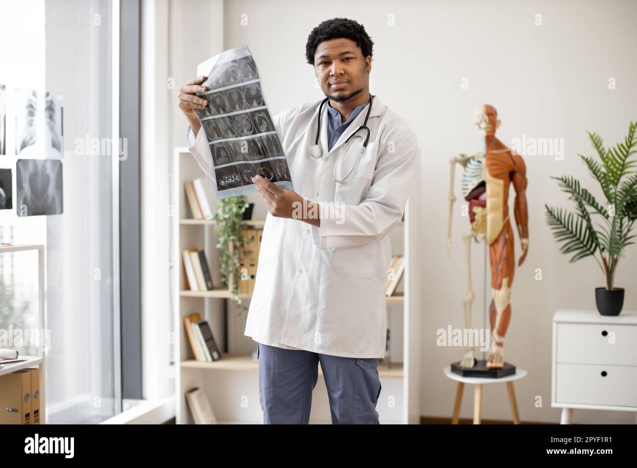 Serious multiracial adult wearing white coat and stethoscope studying CT brain scans while working in doctor's office interior. Mindful healthcare practitioner deciding on correct treatment plan. Stock Photo
