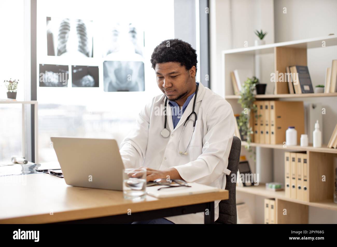 Mindful multicultural adult in white coat with stethoscope using portable computer in doctor's workplace. Health professional developing treatment pla Stock Photo