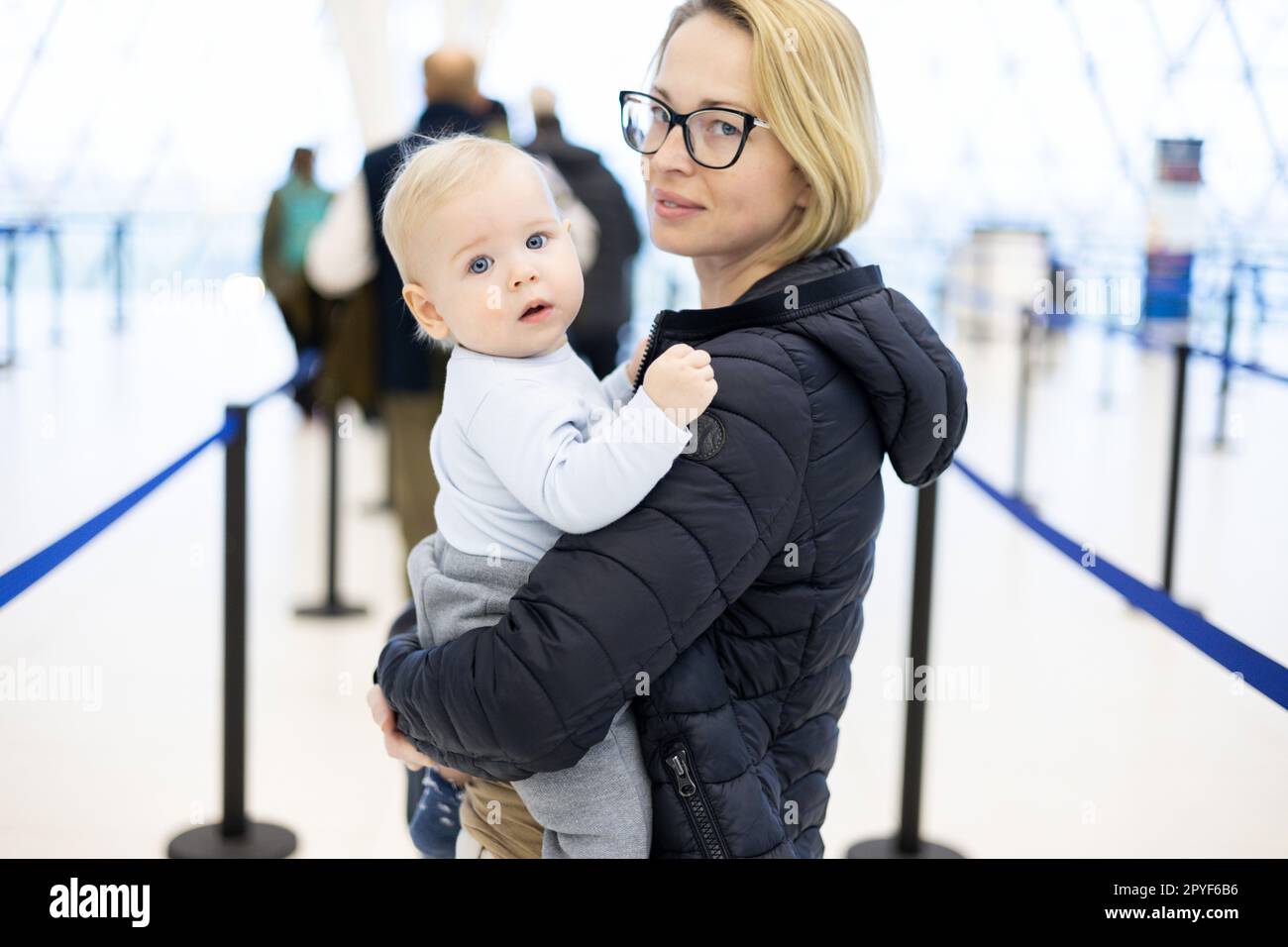 Mother carying his infant baby boy child queuing at airport terminal in passport control line at immigrations departure before moving to boarding gates to board an airplane. Travel with baby concept. Stock Photo