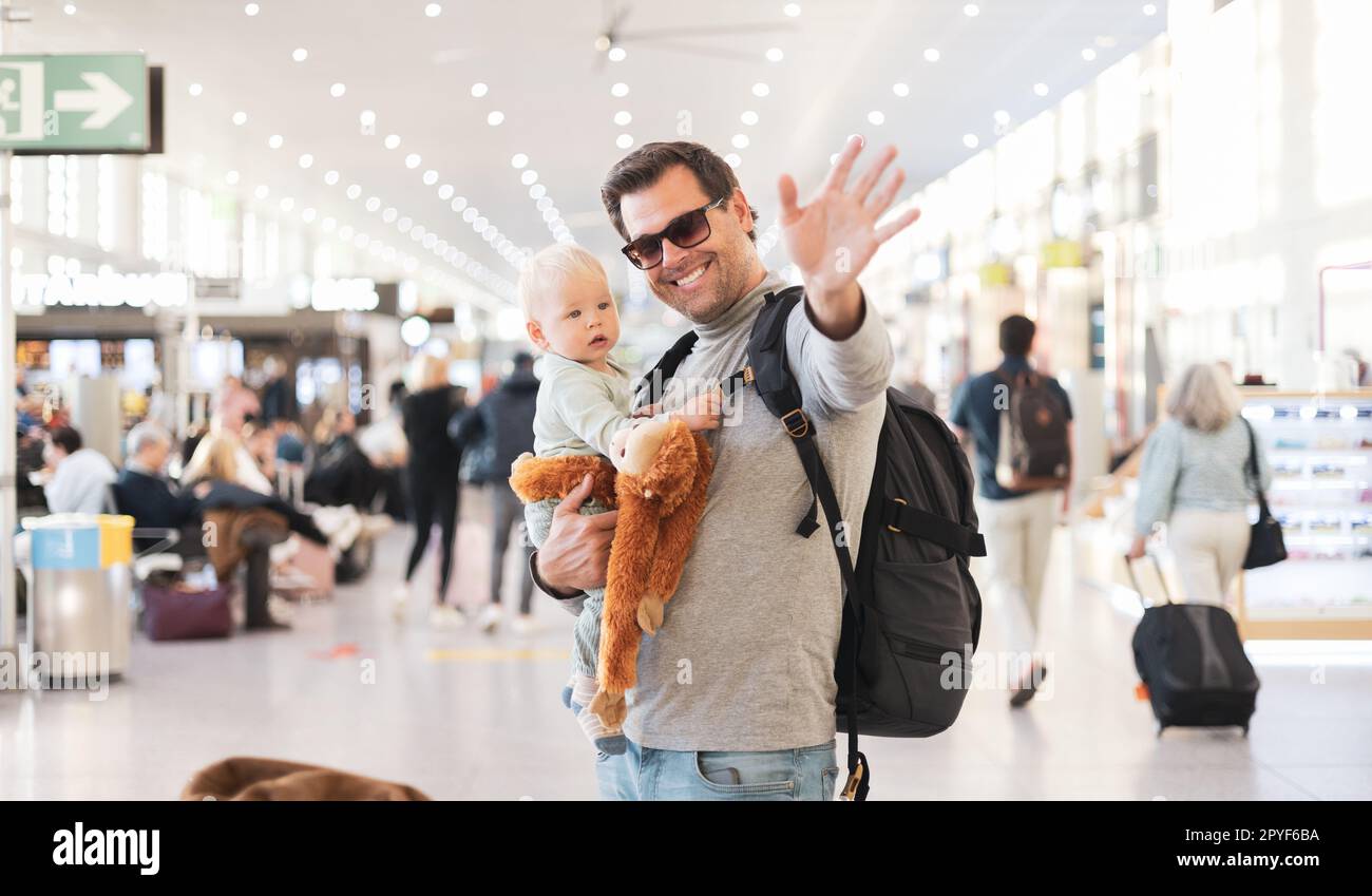 Father traveling with child, holding his infant baby boy at airport terminal waiting to board a plane waving goodby. Travel with kids concept. Stock Photo