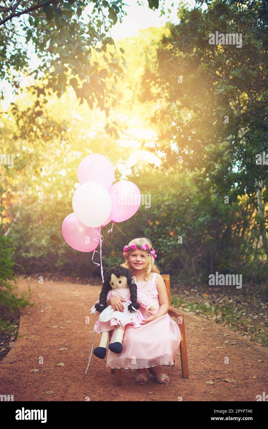 Dolls are a girls best friend. a happy little girl sitting and waiting with a doll and balloons in the middle of a dirt road. Stock Photo