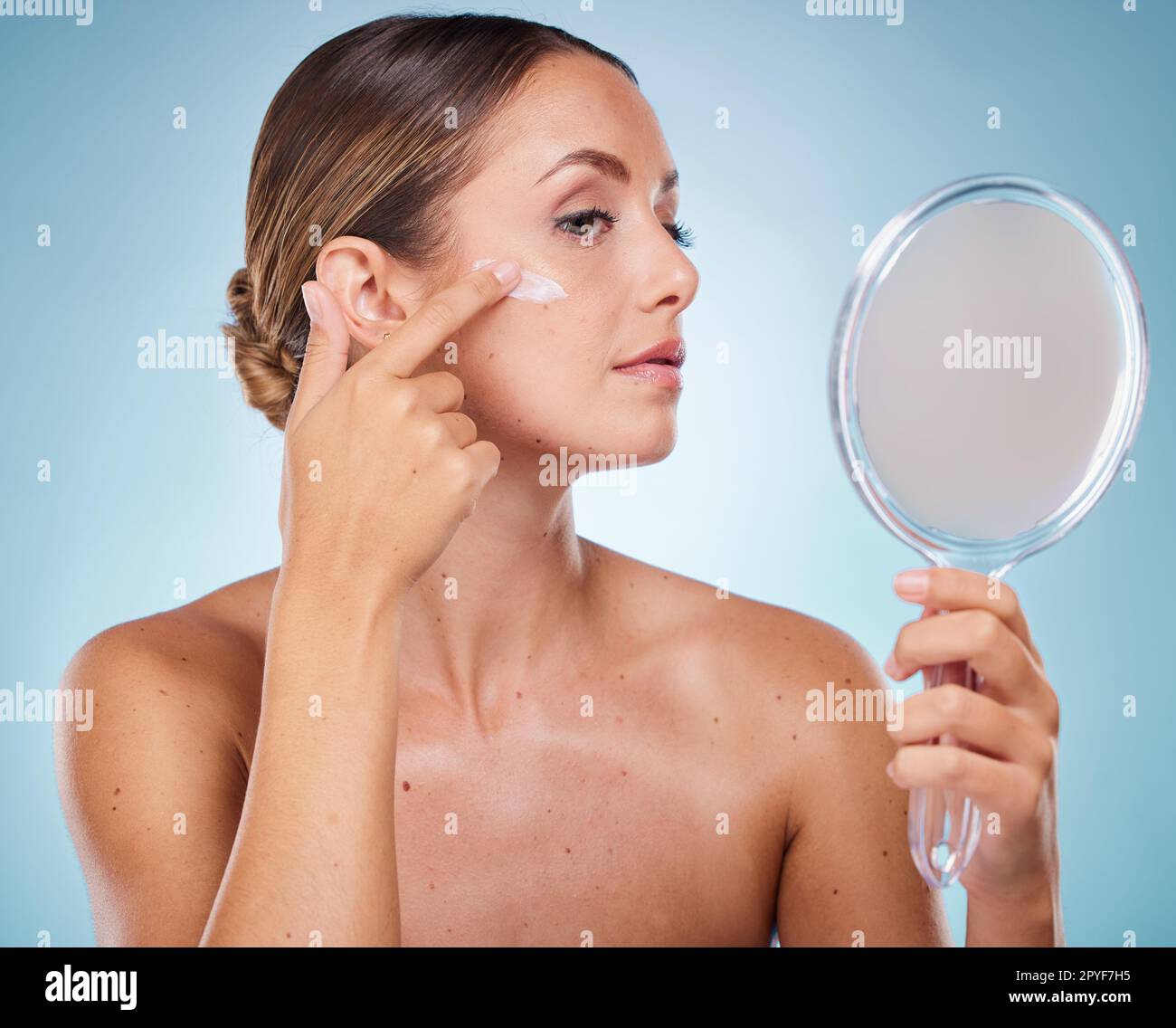Woman, face and cream with hand mirror for beauty wellness, skincare dermatology and cosmetics moisturizer in blue background studio. Model, apply sunscreen and makeup facial product for self care Stock Photo
