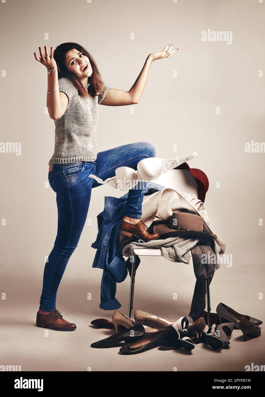 All these clothes and still nothing to wear. Studio shot of a young woman choosing clothing piled on a chair against a brown background. Stock Photo