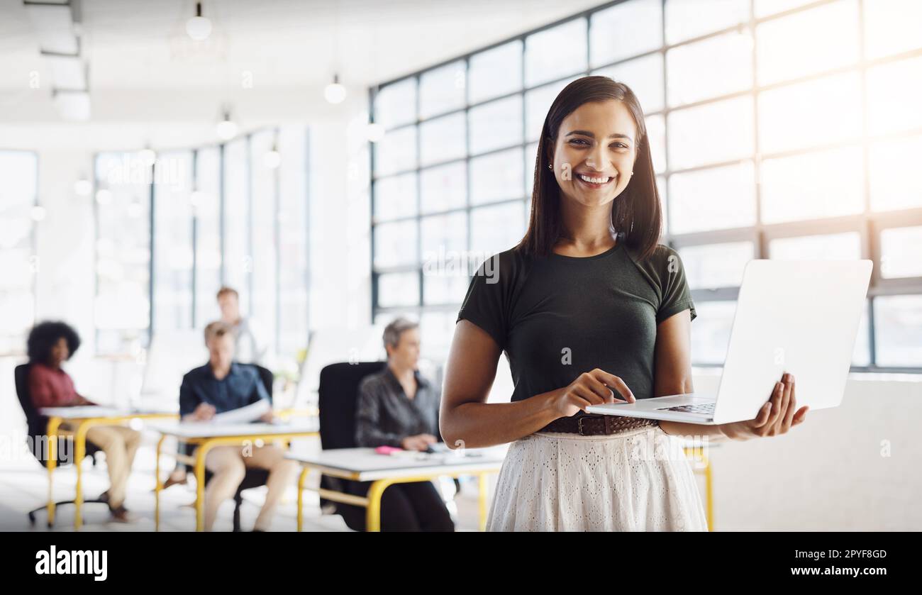 Always happy to help in bringing your ideas to life. Portrait of a happy designer posing in the office while her colleagues work in the background. Stock Photo