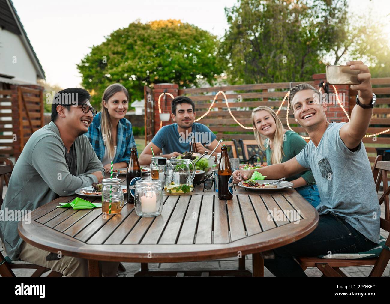 Everyone get closer for the photo. a group of young friends getting closer for a self portrait while being seated outside around a table. Stock Photo