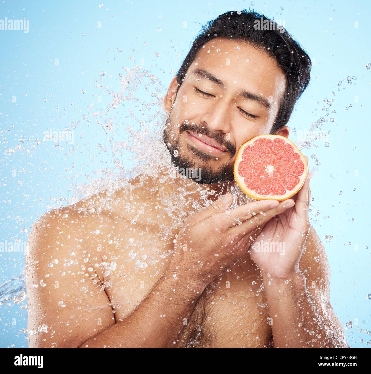 Face, water and grapefruit with a man model in studio on a blue background for hygiene or natural hydration. Skincare, beauty or fruit with a handsome young male wet from a water splash in the shower Stock Photo