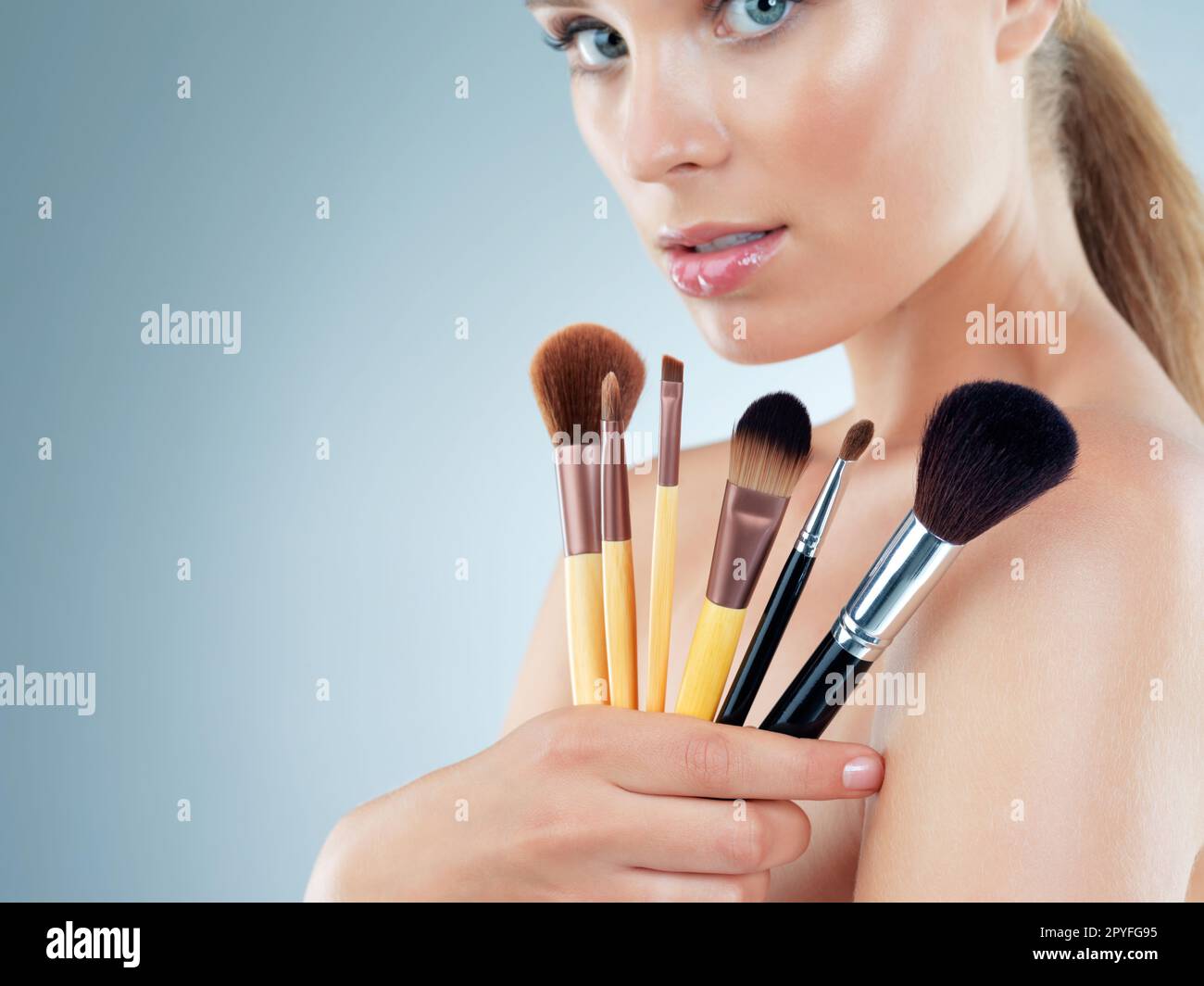 Pick a brush, any brush. Studio portrait of a beautiful young woman posing with makeup brushes against a blue background. Stock Photo