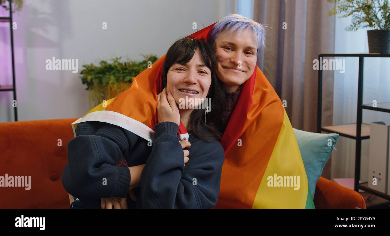 Homosexual lesbian couple sitting at home room couch next to each other,  holding LGBT flag. Smiling