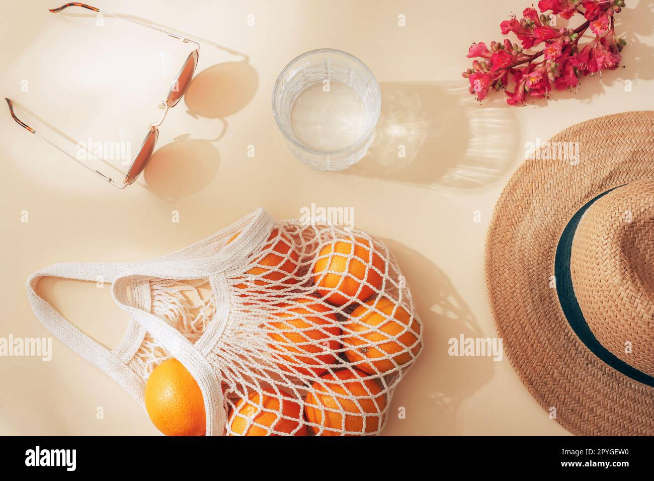 Straw hat, sunglasses, glass of water, string bag with oranges and red flower on beige background in sunlight. Summer concept. Top view, flat lay. Stock Photo