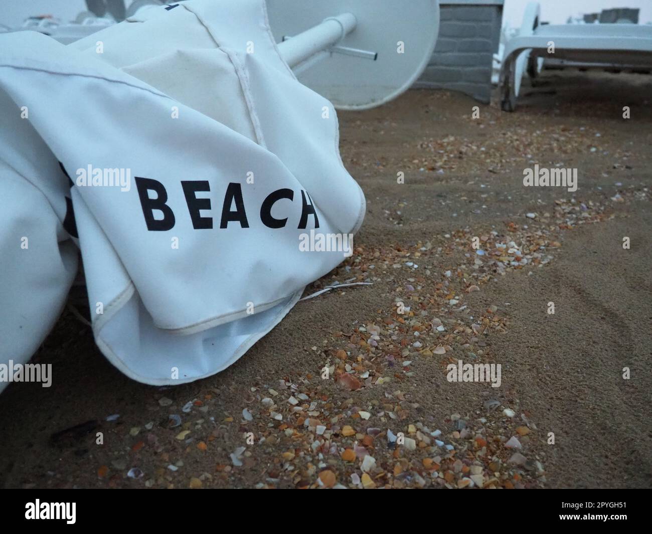 A beach umbrella lay folded on a sandy beach after bad weather. Storm at sea. End of the beach season due to typhoon and cyclone. Loungers, umbrellas and hotel furniture. Beach word written on white Stock Photo