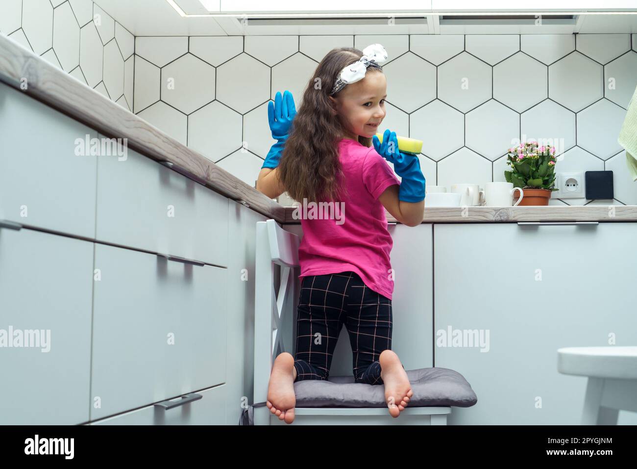 Happy little girl in protective gloves wash up dishes with sponge in kitchen sink sitting on chair. Home cleaning. Stock Photo