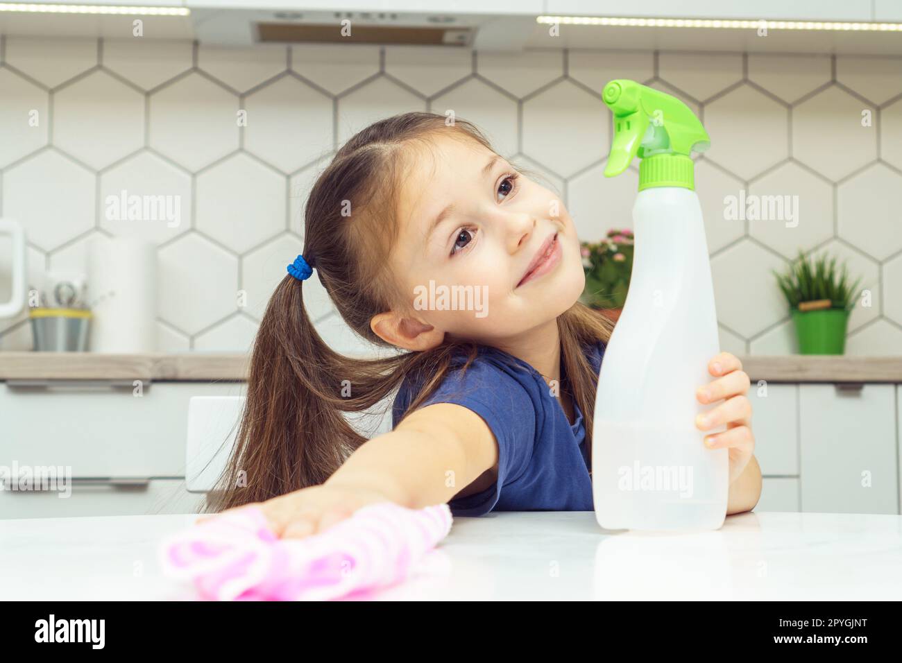 Nice little girl with detergent sprayer and household rag wipe table. Portrait of happy child tidying up kitchen. Stock Photo