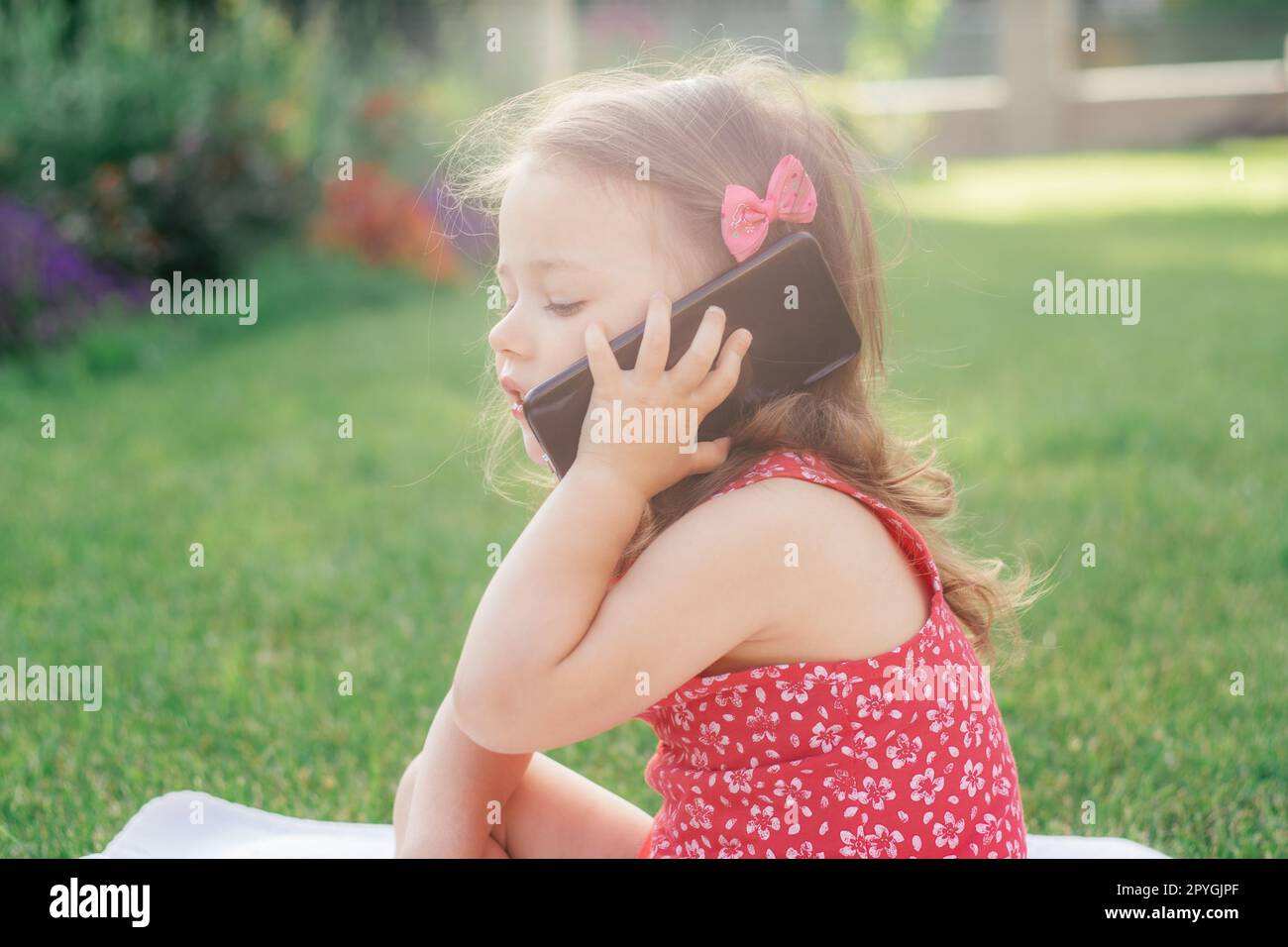 Portrait of little 3-4 girl in red sitting on blanket on green grass and speaking on cell phone. Children using gadgets Stock Photo