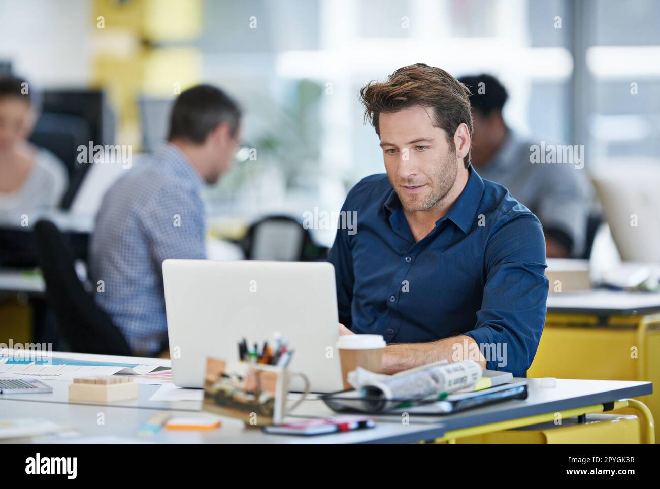 Wrapping his head around the issue. Portrait of a designer sitting at his desk working on a laptop with colleagues in the background. Stock Photo