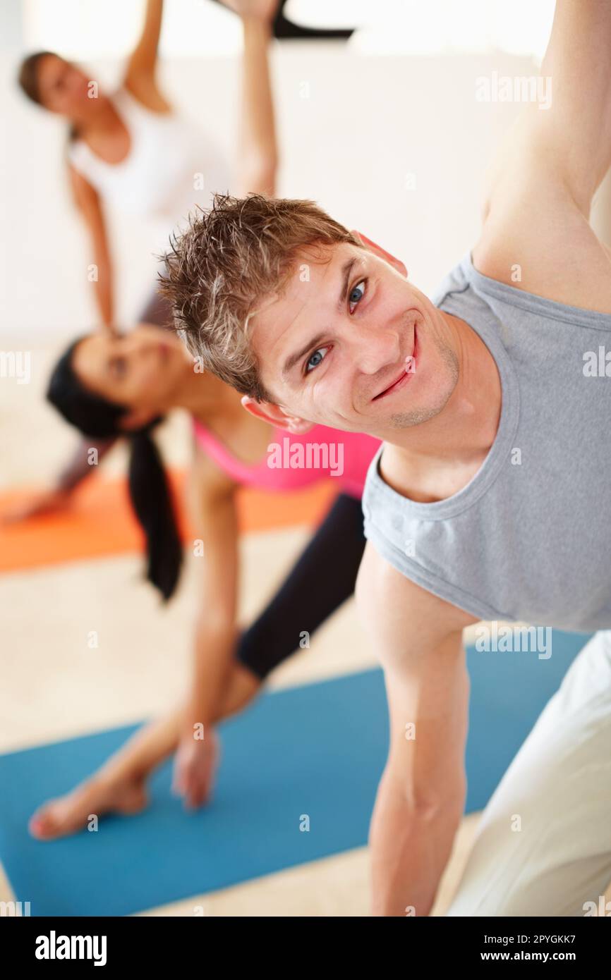 Putting some macho into yoga. A young man smiling at the camera while doing a stretch in yoga class. Stock Photo
