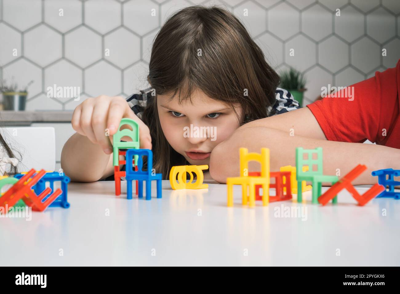 Thoughtful, focused little girl building and holding small green chair toy near many colorful chair toys on white table Stock Photo