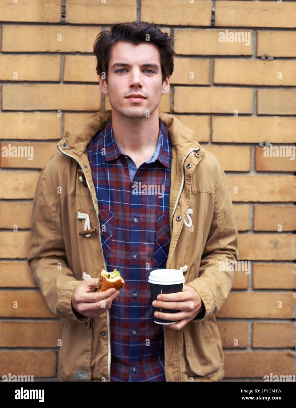 Food for his mind, body and soul. a young man standing outdoors with a half eaten burger and take away coffee. Stock Photo