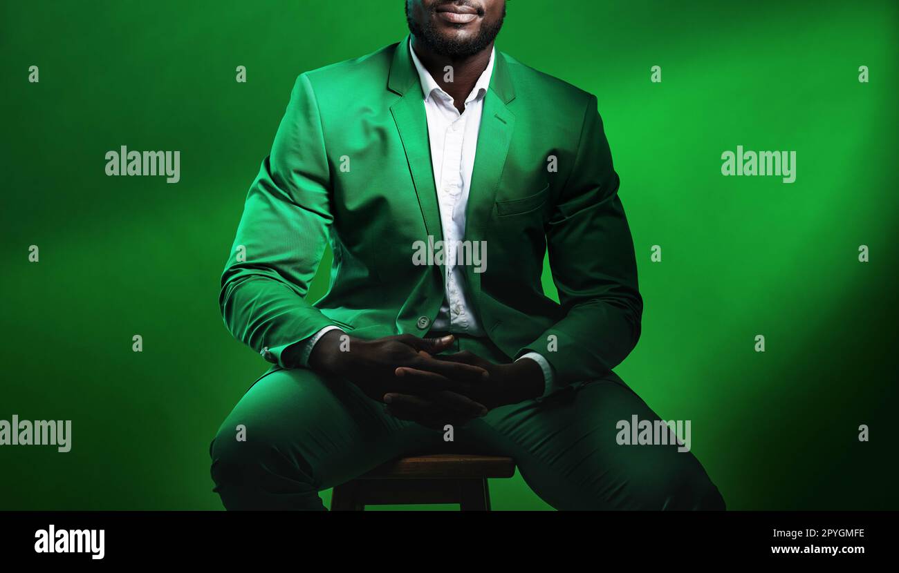Fashion, suit and black man model with a formal outfit sitting on a chair while posing in a studio. Elegant, trendy and African male in luxury, stylish and fancy clothes isolated by green background. Stock Photo