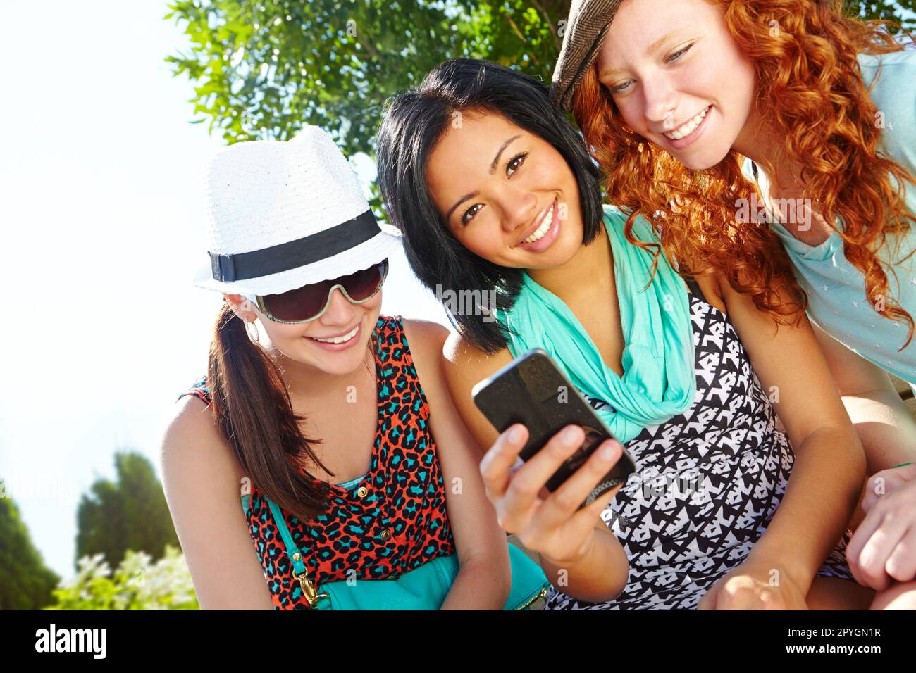 Feeling social. Three teenage girls sitting outside with a cellphone smiling at a text message. Stock Photo