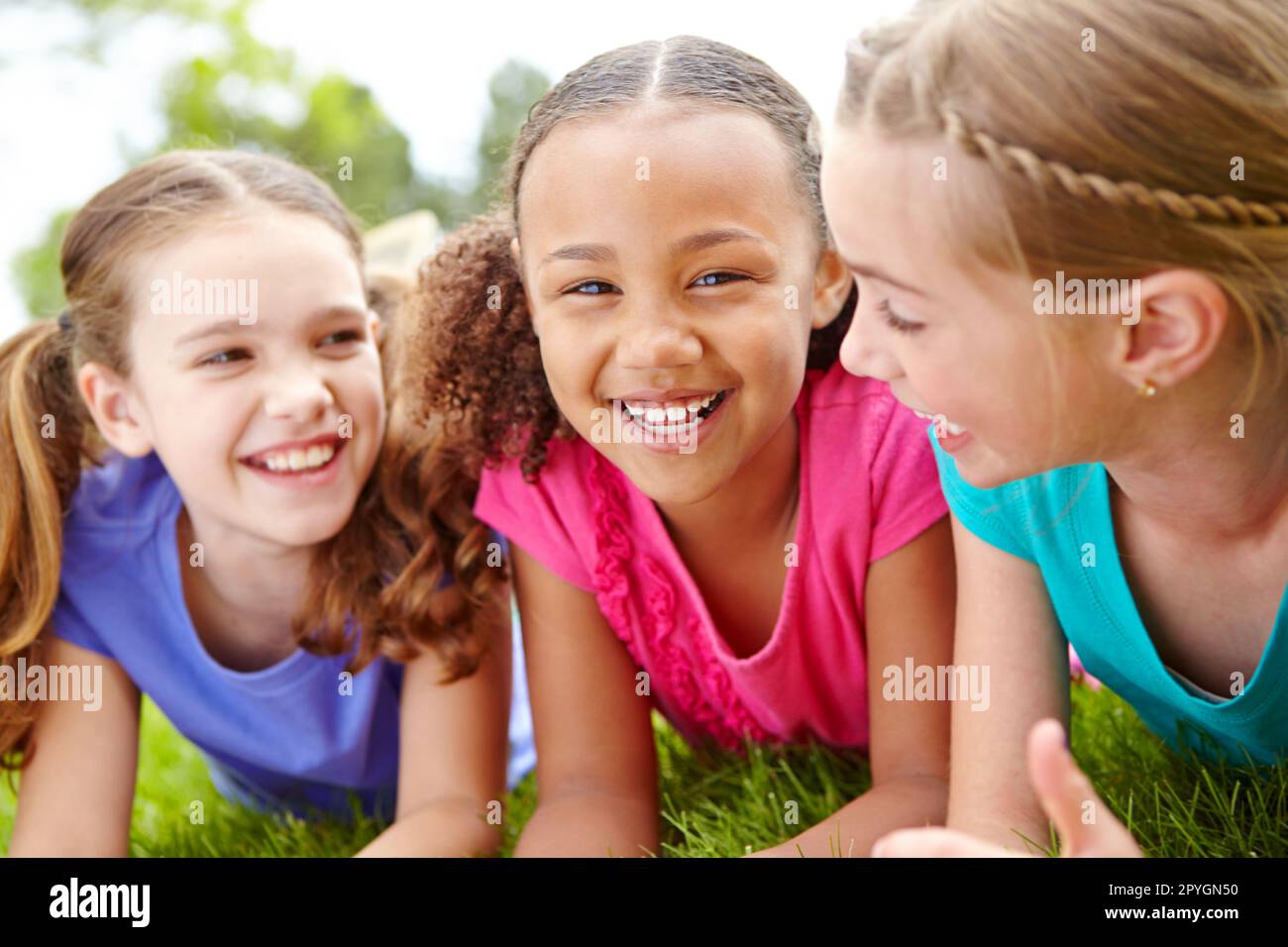 Theyre such a terrific trio. Three multi-ethnic young girls lying on the grass in a park smiling at the camera. Stock Photo