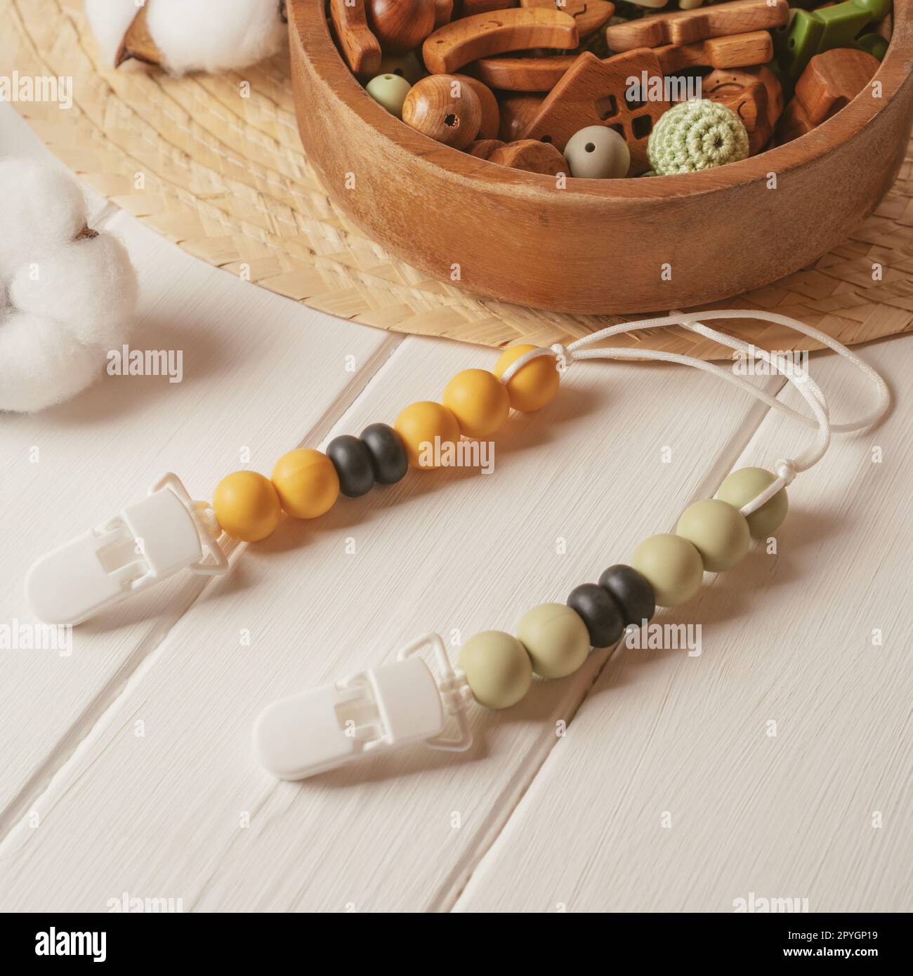 White plastic clothespin with white, black and orange wooden beads on white laces for baby pacifiers near wooden bowl. Stock Photo