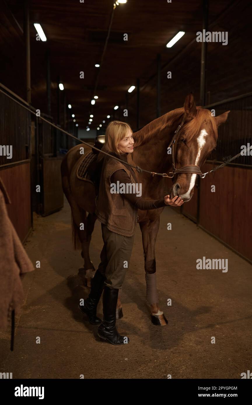 Horsewoman petting horse stroking animal muzzle over stable interior Stock Photo
