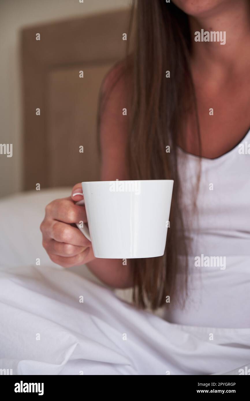 White cup in the hands of a young woman sitting on the bed in the bedroom in the morning. Stock Photo