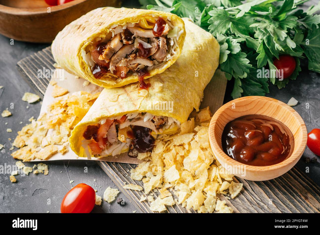 Crispy shawarma with chips, barbeque sauce, chicken in pita bread on wooden dark surface with cherry tomatoes, parsley Stock Photo