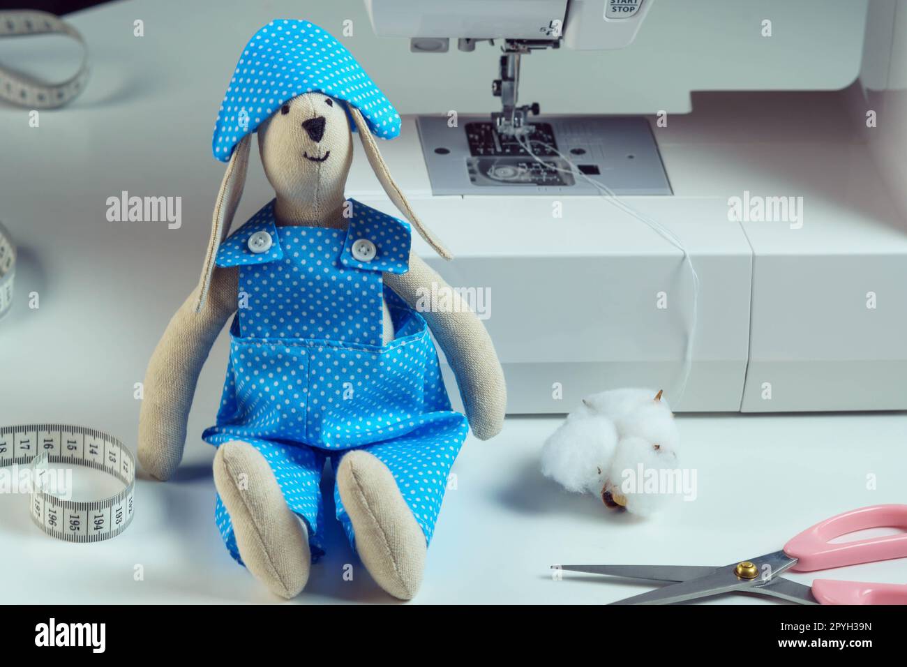 Funny smiling sewed stuffed rabbit toy in blue overalls, cocked hat, sit near cotton, scissors, next to sewing machine Stock Photo