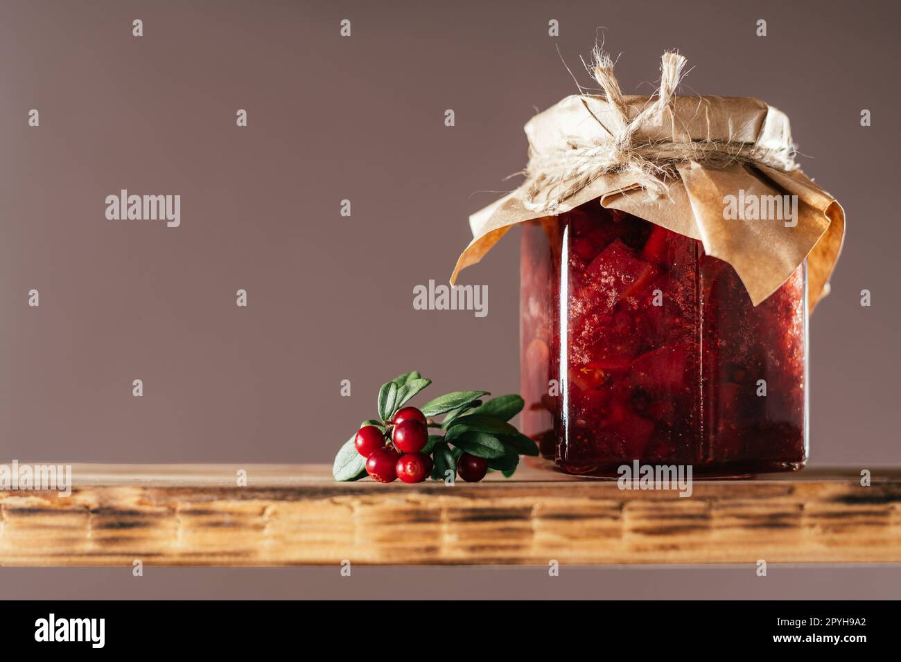 Jar of lingonberry and pear jam with craft paper on lid on wooden shelf next to fresh lingonberries on brown background Stock Photo