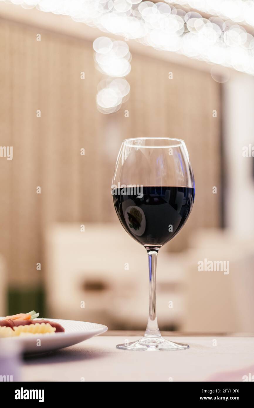 Vertical photo of dark dry wine vine glass on blurred background. Light restaurant, cafe indoor. Food plate. Close up Stock Photo