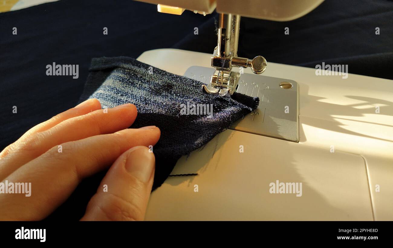Sewing process on a modern sewing machine, close-up. Sewing machine foot with a needle. Dark blue fabric is trimmed with double wrapping of the fabric edge. Natural sunlight Stock Photo