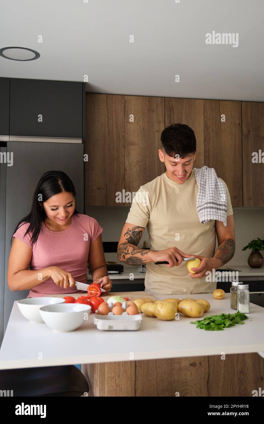 Two young latin people peeling potatoes and cutting tomatoes at kitchen. Stock Photo