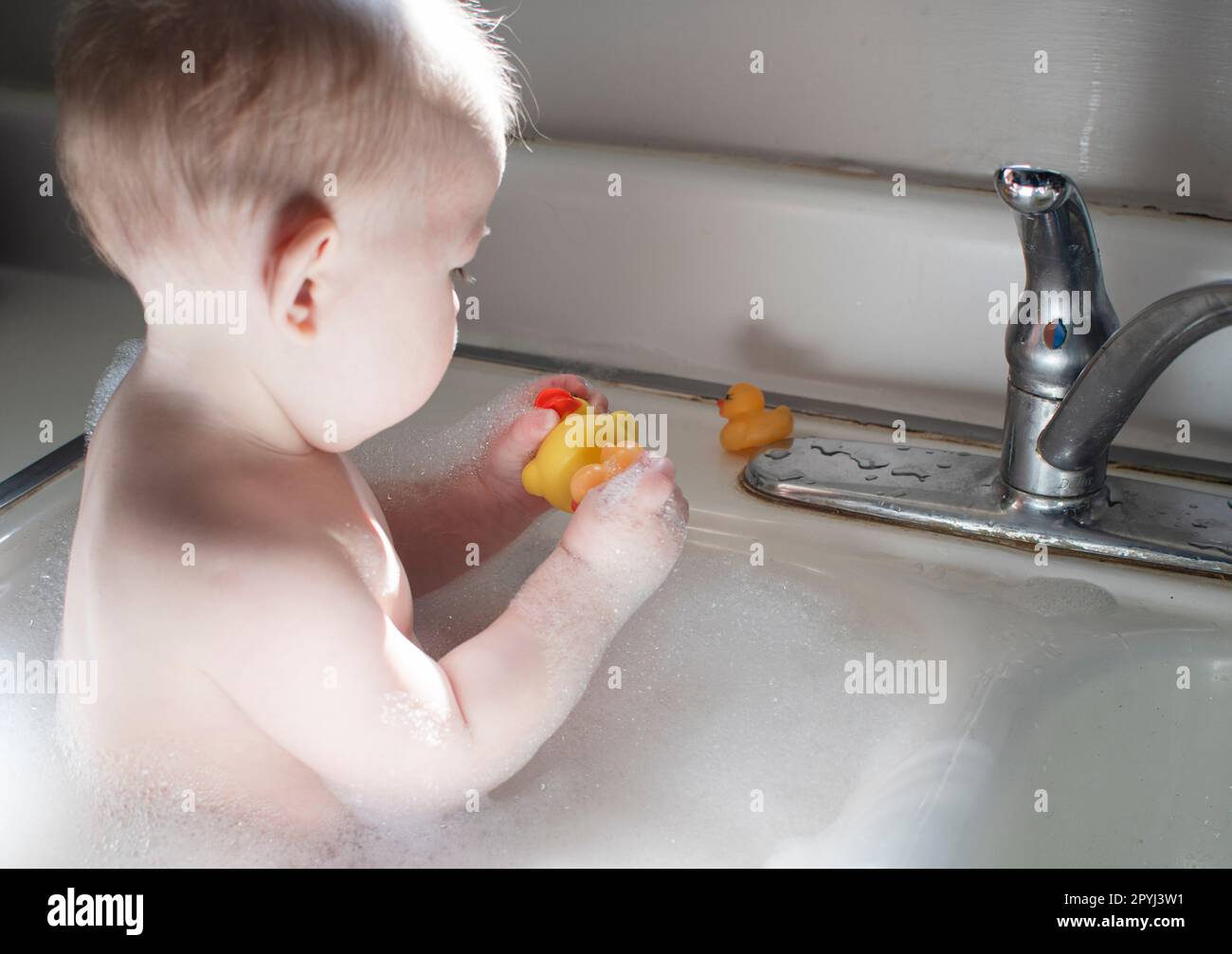 Baby taking bath in the kitchen sink. Child playing with foam and soap