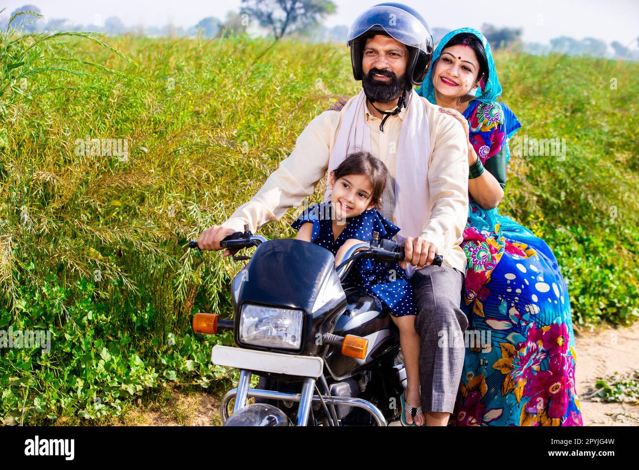 Happy young rural Indian family wearing safety helmet riding on motorcycle in village agricultural field. Mode of transportation, Copy space Stock Photo