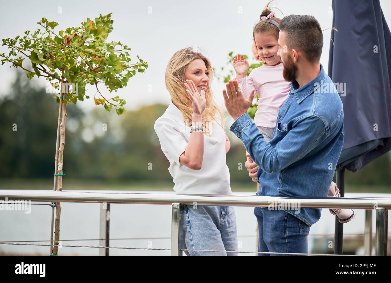 Cheerful woman, man and little girl standing near green tree and slapping hands giving high five outdoors. Happy man holding daughter and smiling while celebrating success with wife and child. Stock Photo