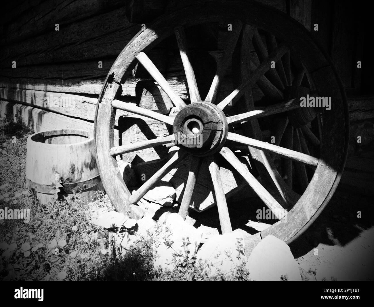 Wooden wheel from a cart. Decorative wheels for decorating lawns, exteriors and rustic interiors. Round homemade wheel against the wall. Retro or vintage style. Countryside life. Black and white Stock Photo