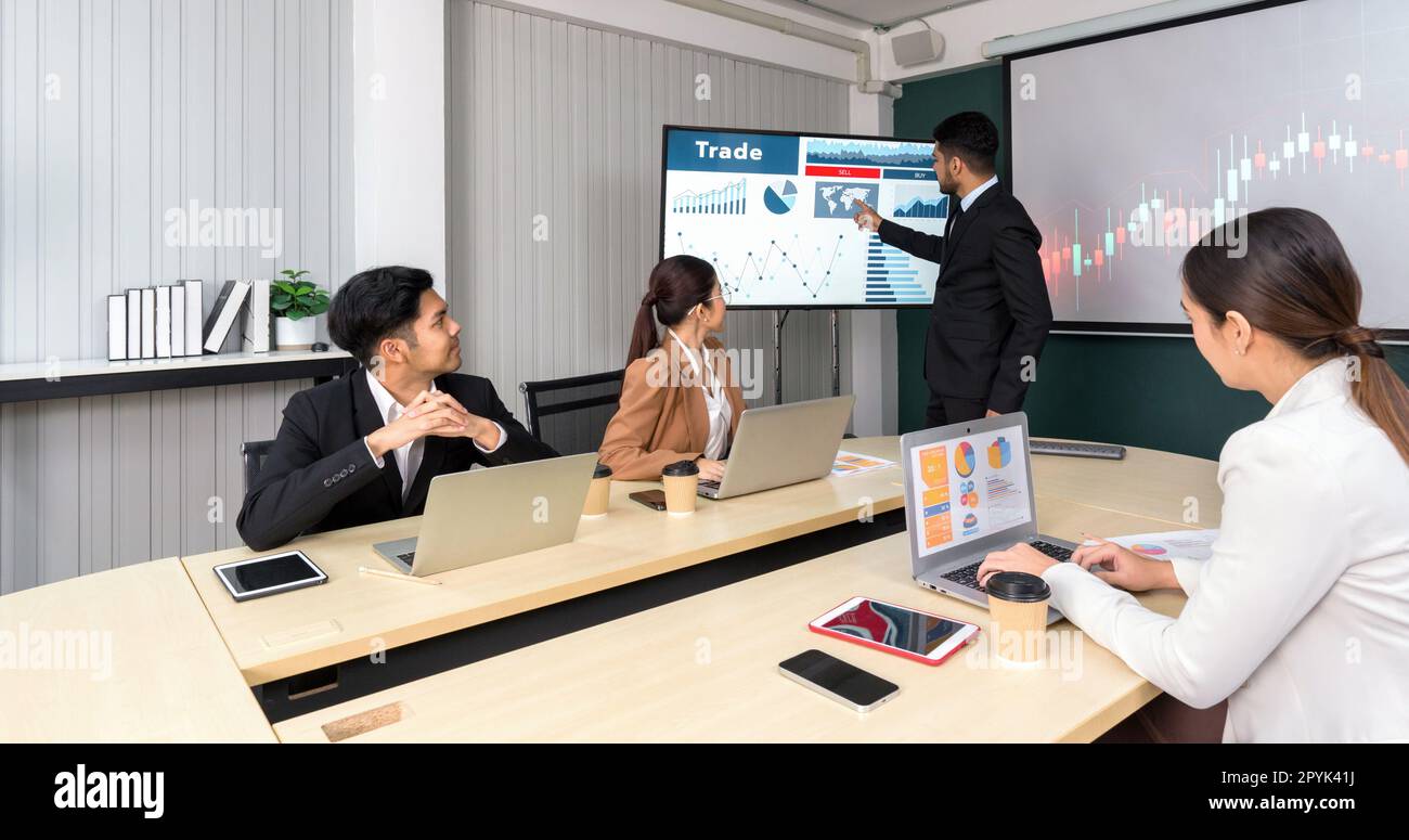 Businessman in black suit pointing at global map on large digital monitor. Business executives team meeting in modern office with laptop computer, tablet, mobile phone and coffee on table. Stock Photo