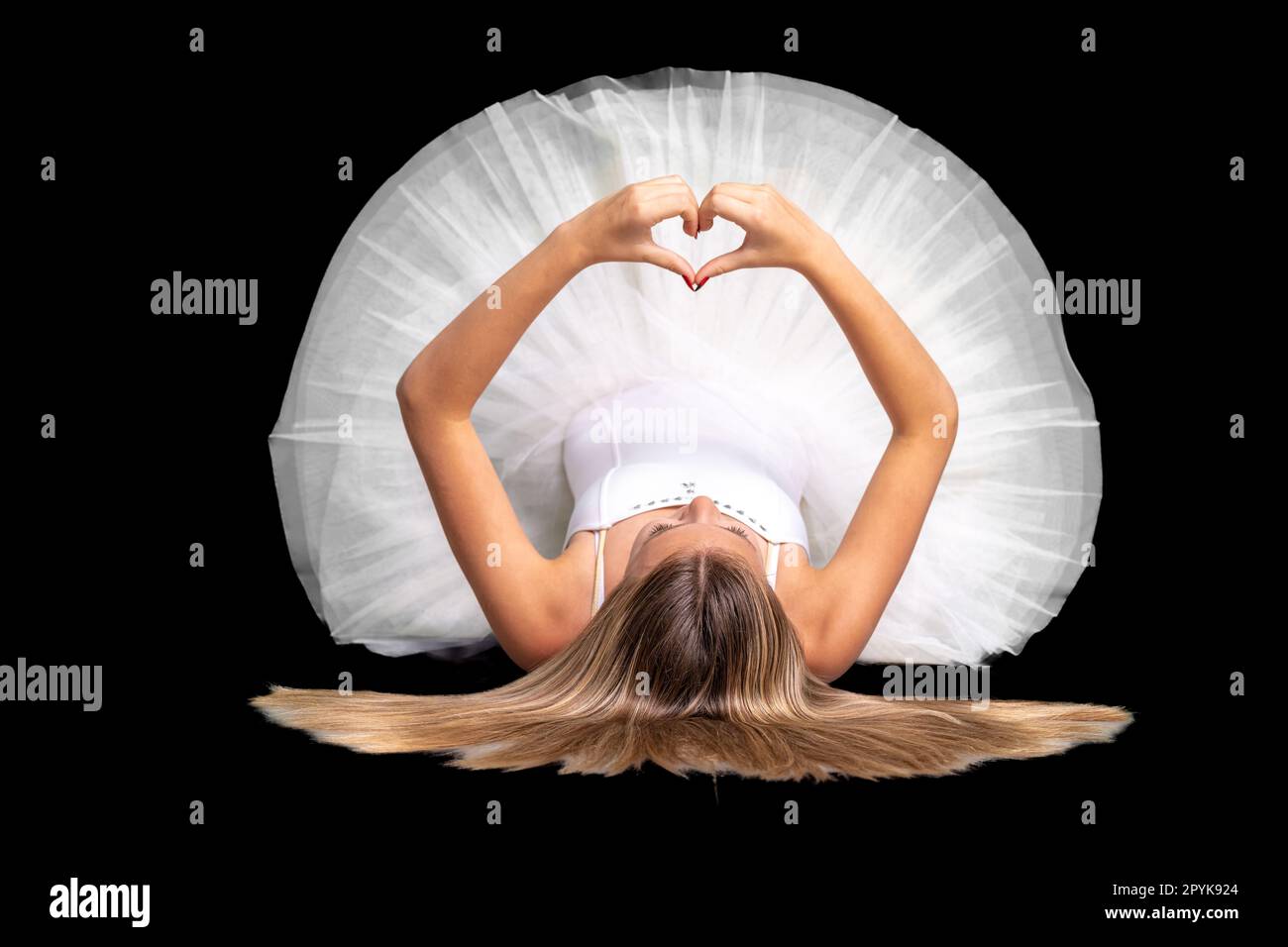 ballerina in a white dress lies on the ground and shows a heart with her hands, portrait on a black background Stock Photo