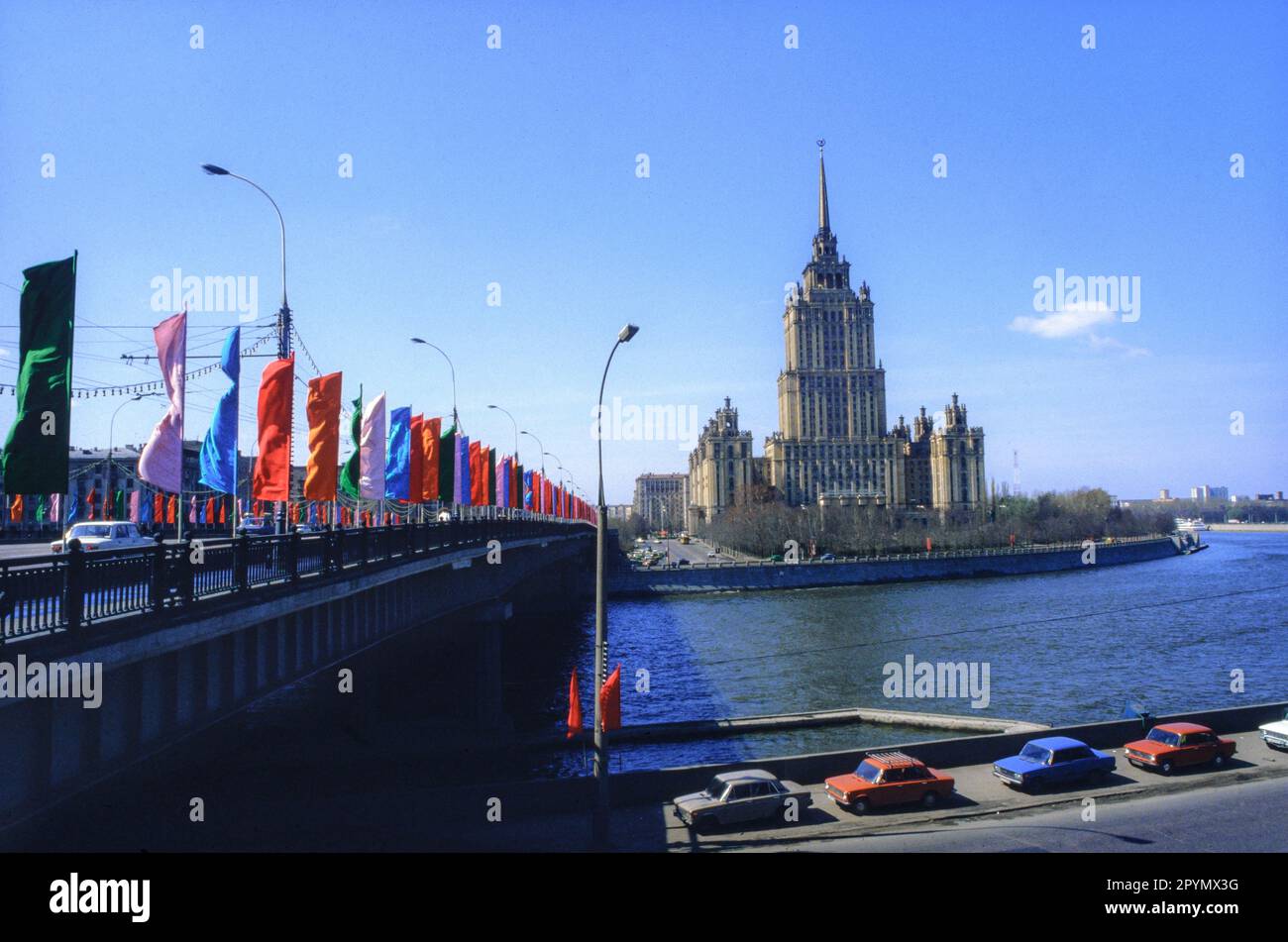 1988: Flags fly on the Kutuzovsky Bridge over the Moskva River in front of the Hotel Ukraina in preparation for the May Day celebrations. The Baroque 'wedding cake' style building is one of the so-called 'Seven Sisters' commissioned by Joseph Stalin as part of a plan to modernize Moscow immediately following World War II. Construction began in 1953 and the Hotel Ukraina opened in 1957. Stock Photo