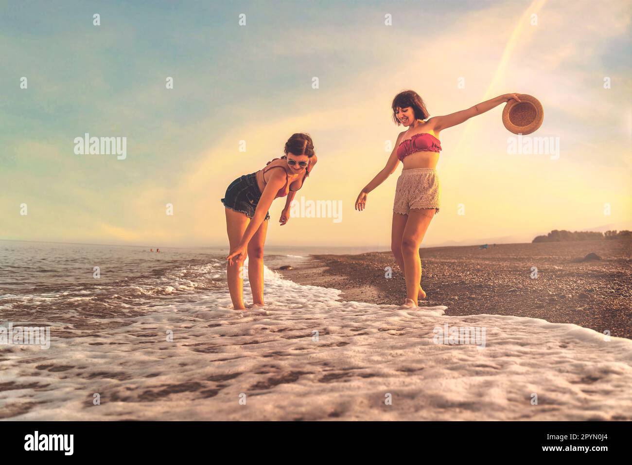 Two young Caucasian young women in their twenties, wearing shorts and bikinis, playfully splash water on their feet at the beach during sunset. One ho Stock Photo