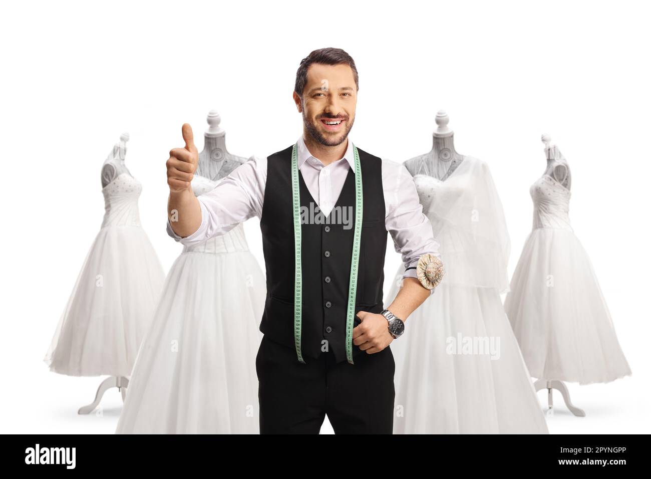 Young tailor with a measuring tape smiling at camera and showing thumbs up in front of bridal gowns isolated on white background Stock Photo