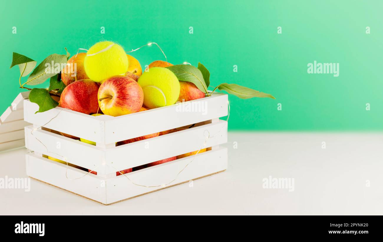 Tennis composition with yellow tennis balls, apples in a white wooden box with lightening on white table on green background. Tennis competition. Conc Stock Photo