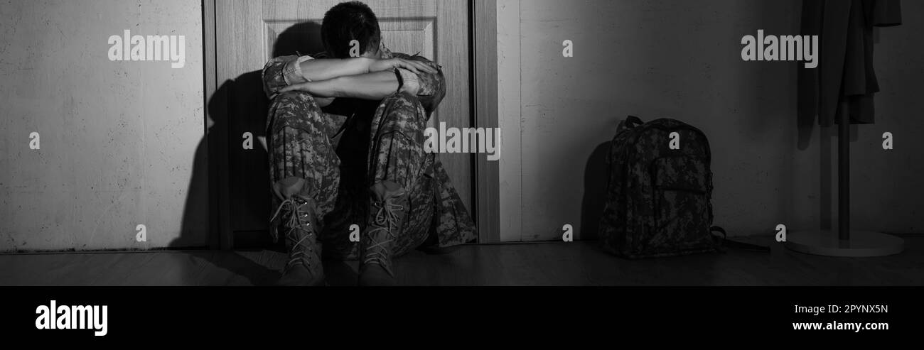 Black and white photo of depressed soldier sitting near backpack and door in hallway at home, banner,stock image Stock Photo