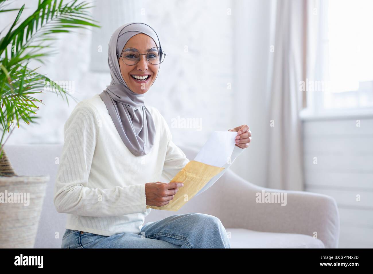 Happy young muslim woman in hijab sitting on sofa at home and opening letter from envelope. Received good news. Smiling, she looks at the camera. Stock Photo