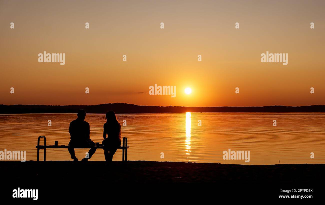 A peaceful sunset on the lake shore. Silhouettes of a man and a woman on a bench. Indefinable people Stock Photo