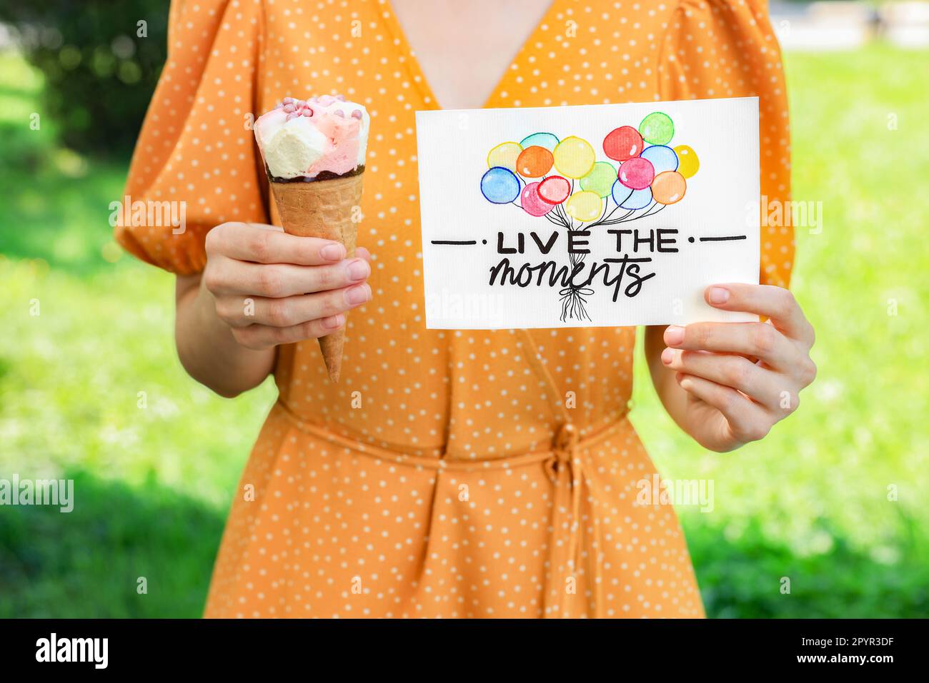 Woman holding card with phrase Live The Moments and ice cream outdoors, closeup Stock Photo