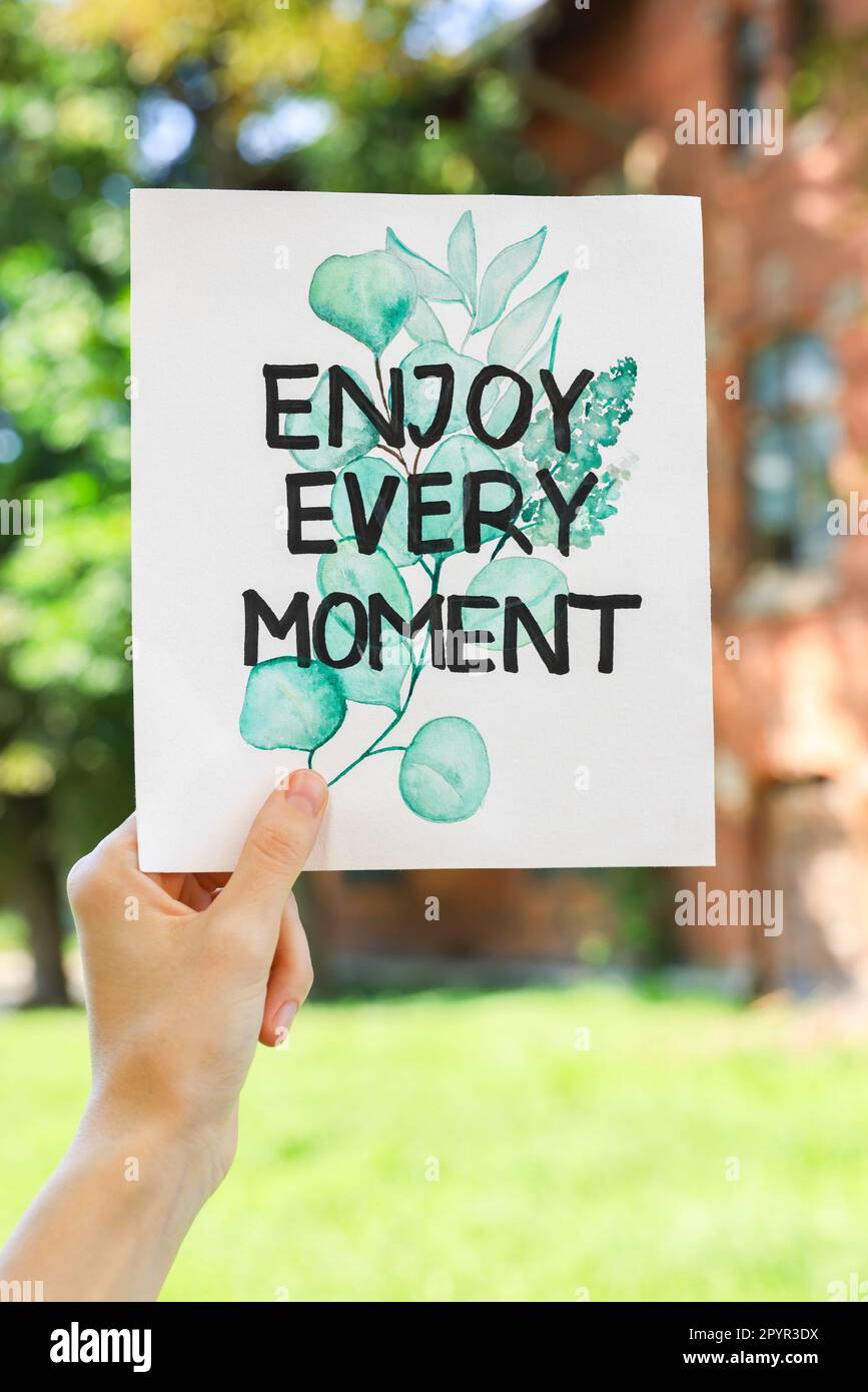Woman holding card with phrase Enjoy Every Moment outdoors, closeup Stock Photo