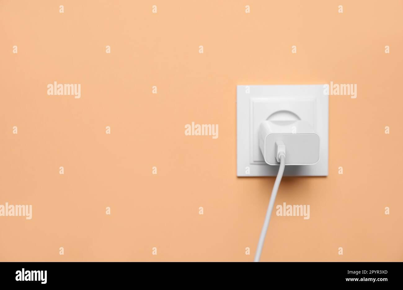 Charger adapter plugged into power socket on pale orange wall, space for text. Electrical supply Stock Photo